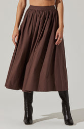 A woman is wearing a high-waisted, full-length brown skirt with pleats, paired with tall black boots.